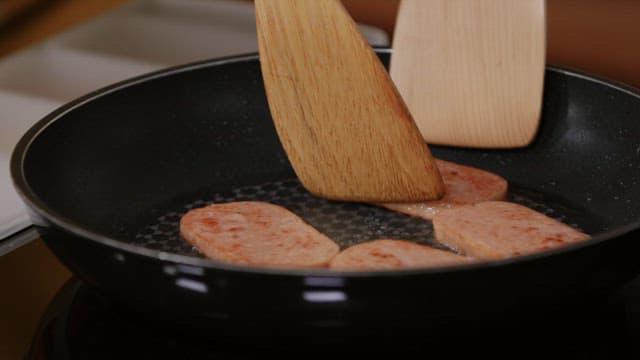 Cooking ham slices on a hot frying pan using wooden spatulas