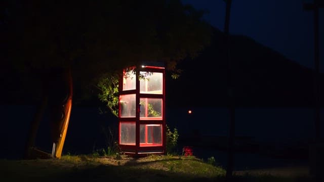 At night, a red phone booth is illuminated in a dark park