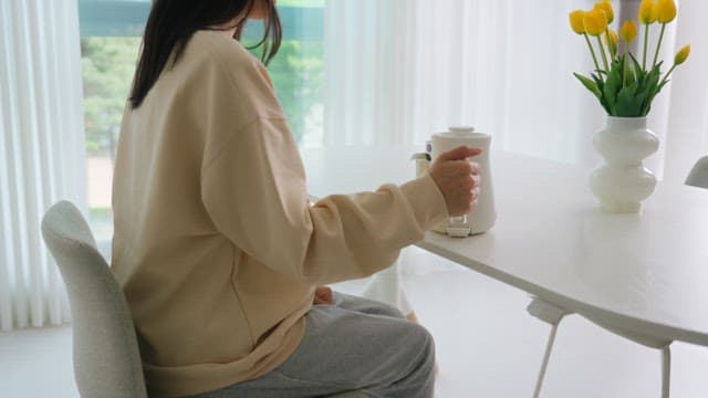 Woman pouring water into cup with coffee pot on table in bright living room