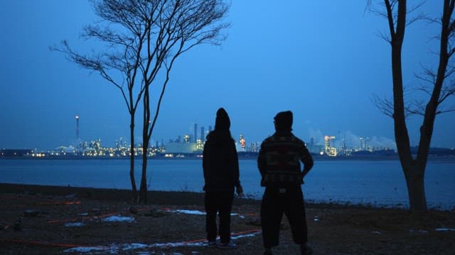 Two people looking at a lit industrial city from the beach under a blue sky at dusk