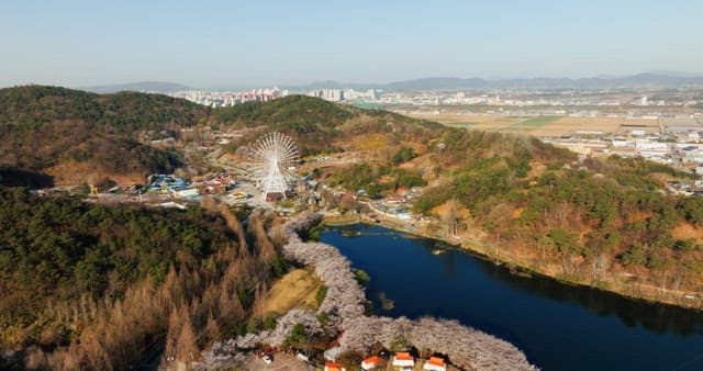 View of a park with a ferris wheel next to a serene lake surrounded by lush greenery and hill