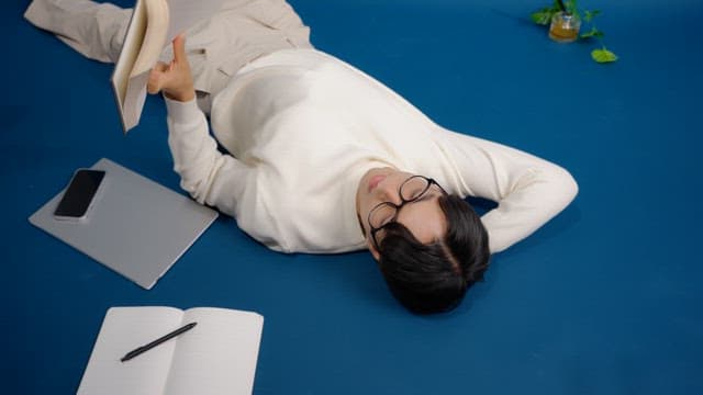 Man lying on the floor reading a book with laptop, notebook, and phone nearby