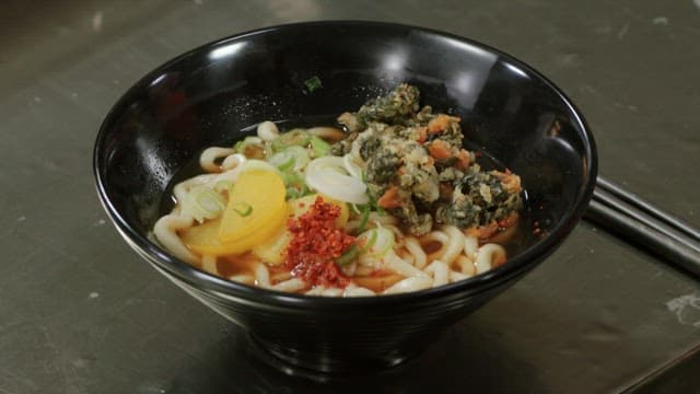 Udon noodle soup in black bowl with colorful garnishes.