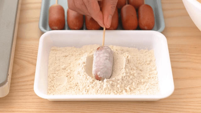 Scene where a sausage stuck on a toothpick is coated with white powder