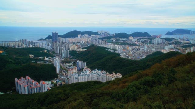 Panoramic view of a coastal city surrounded by green mountains under a cloudy sky