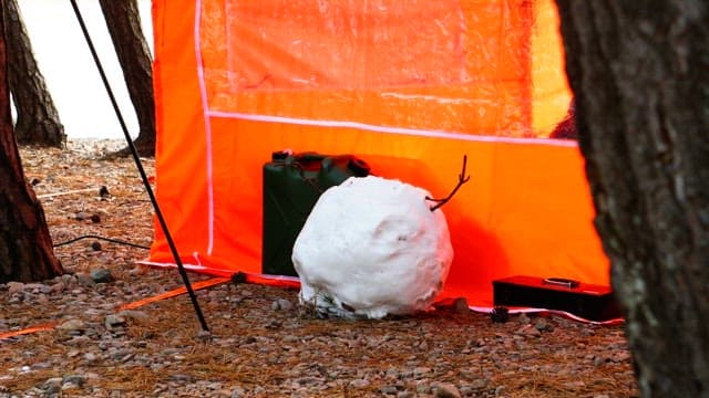 Person assembling a snowman next to an orange tent in a wooded area