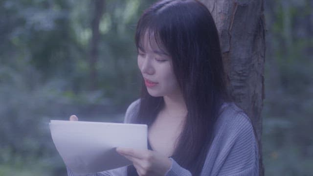 Woman with long hair writing on a paper while leaning against a tree in the forest