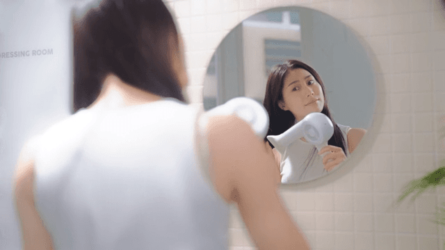 Woman drying her hair with a hair dryer in front of a bathroom mirror