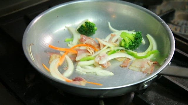 Stir-frying vegetables and bacon in a pan on the stovetop with sparks
