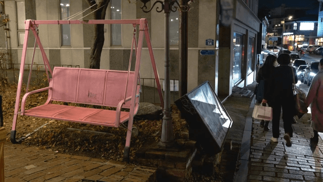 Pedestrians and cars passing by on a street lined with decorative pink swings