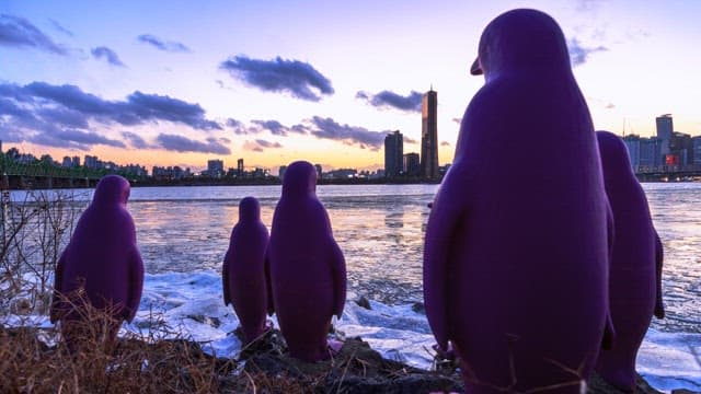 Purple penguin sculptures against the backdrop of a cityscape transitioning from sunset to night