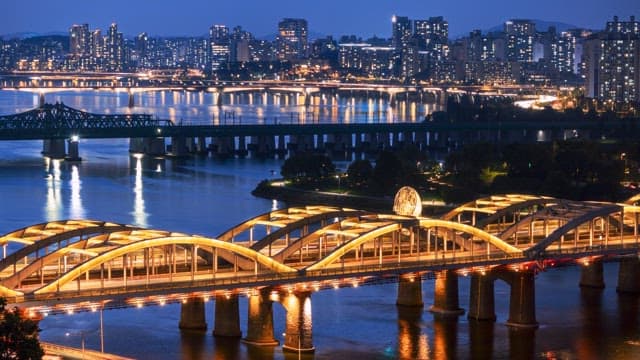 Brightly illuminated bridge crossing a river at night, with a large city skyline in the background