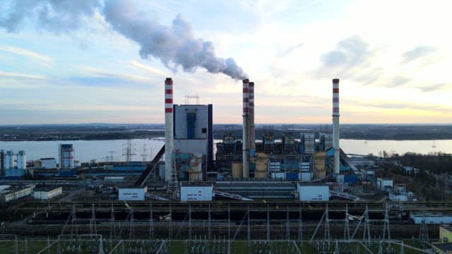 Industrial plant with smoke stacks near a river during sunset.