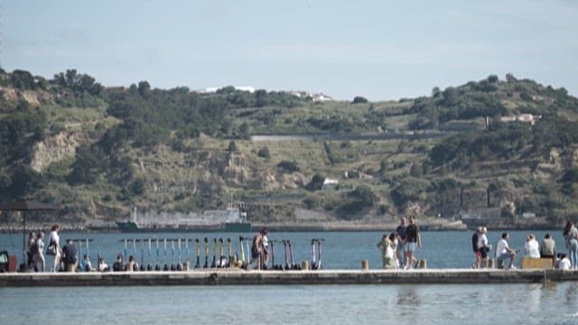 People Enjoying a Sunny Day on a Lakeside Pier with Scenic Hill in the Background