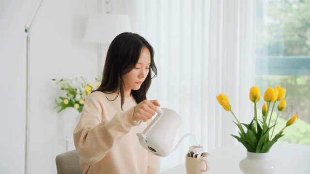 Woman pouring water into cup with coffee pot on table in bright living room