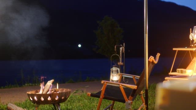 Nighttime campsite with a lit campfire and lantern beside a chair near a table
