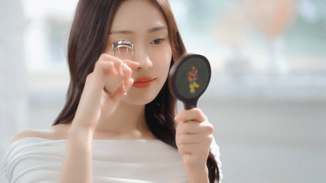Woman using eyelash curler while looking in the mirror in a bright room