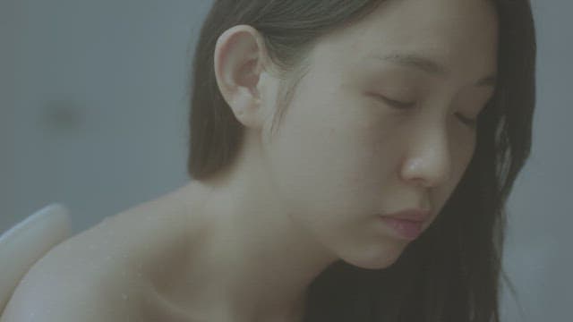 Woman in a bathroom, appears contemplative and slightly melancholic.