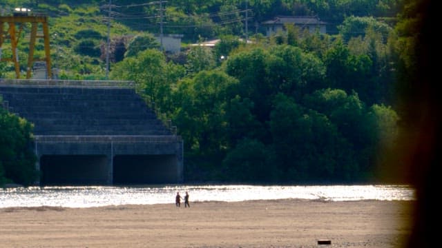 Two people walking hand in hand along a riverside lined with greenery