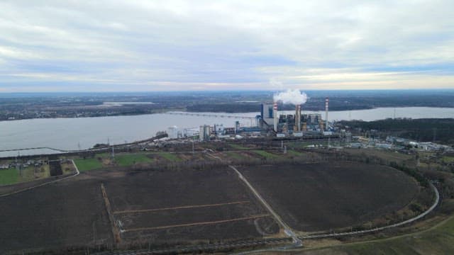 Industrial facility near a large river with smoke stacks and fields