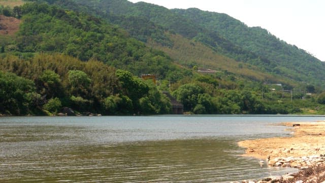View of a serene river flowing beside a lush green hill under clear sky.