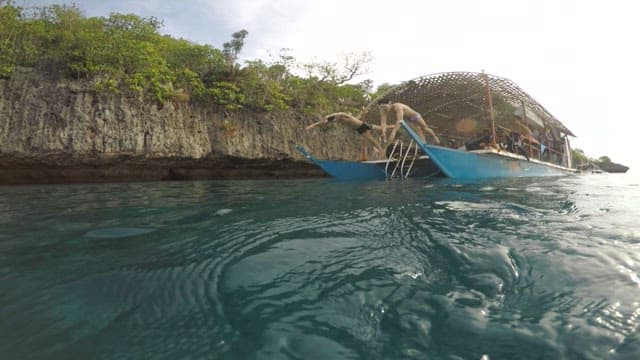 Two men diving off a boat into clear blue water near a scenic rocky and green coastline.