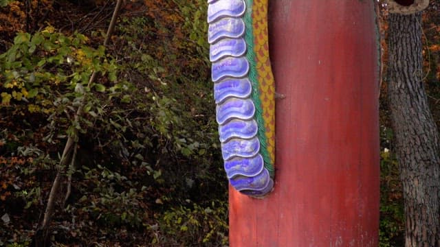 Vibrantly colored dragon sculpture made of wood attached to a red pillar in a forest.